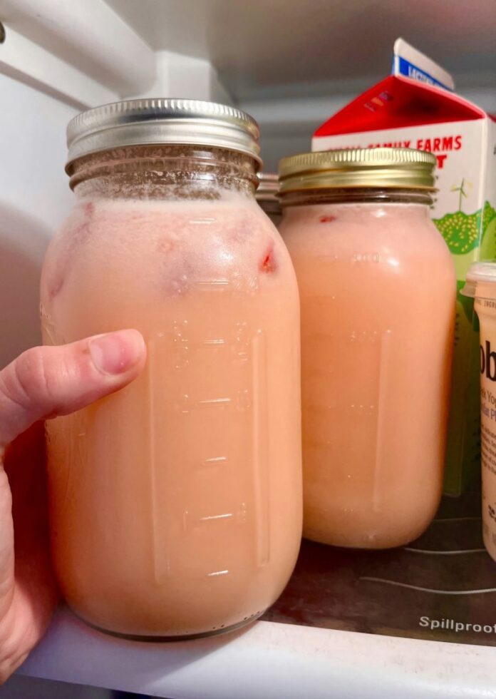 Alright, let's analyze the image you provided! Alt Text: Two glass mason jars filled with a light pink Strawberries Cream Lemonade inside a refrigerator. The jar on the left is being held by a hand.