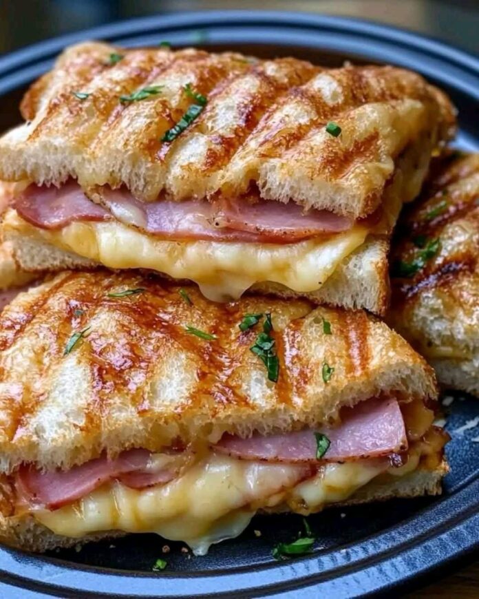 Close-up of two halves of a grilled Hawaiian Ham and Cheese Sandwich stacked on a dark plate. The sandwich has grill marks and is filled with ham and melted cheese. Chopped herbs are sprinkled on top.
