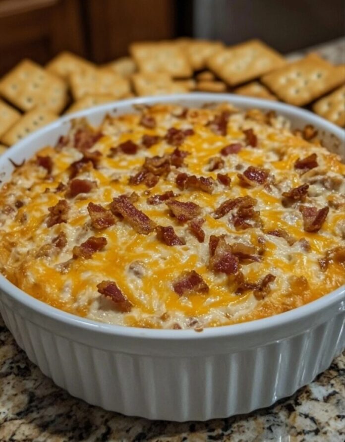 A white, fluted baking dish filled with a creamy dip, topped with melted cheese and crumbled bacon. Square crackers are visible in the blurred background.