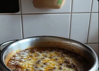 Hearty Ground Beef Dip A white pot filled with a ground beef dip, topped with melted cheese, resting on an orange surface. Tiled wall is visible in the background.