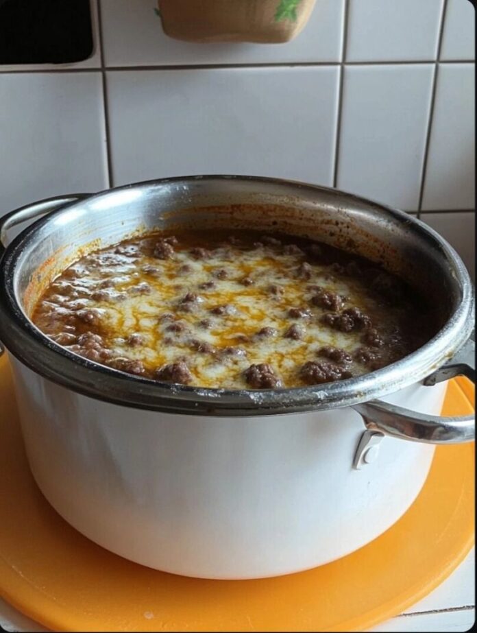 A white pot filled with a ground beef dip, topped with melted cheese, resting on an orange surface. Tiled wall is visible in the background.