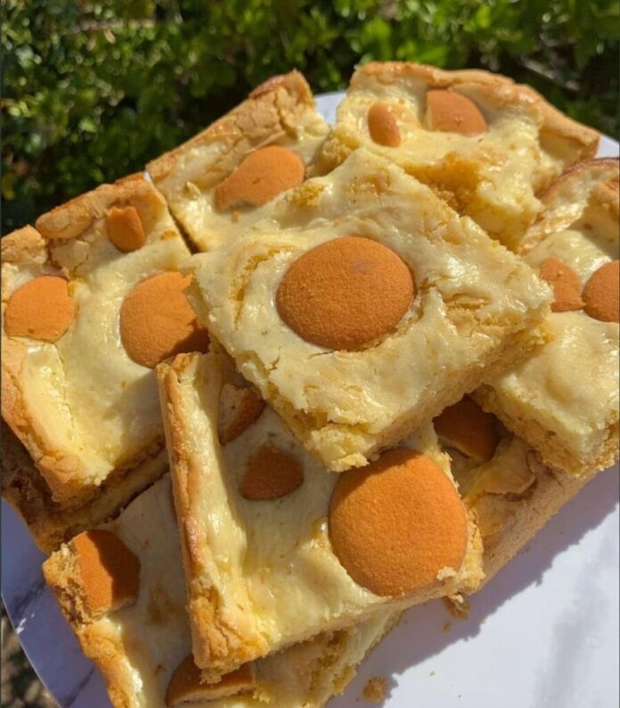 A close-up, overhead shot of several Banana Pudding Brownies cut into squares and stacked on a white plate. Each brownie has a light-colored batter with a circular vanilla wafer cookie pressed into the top. Green foliage is visible in the blurred background.