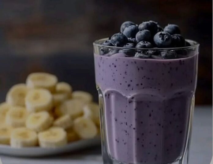 A close-up shot of a glass of Blueberry Banana Smoothie. The smoothie is a purplish-blue color with visible flecks of blueberries. Fresh blueberries are piled on top of the smoothie. A plate of sliced bananas is blurred in the background.