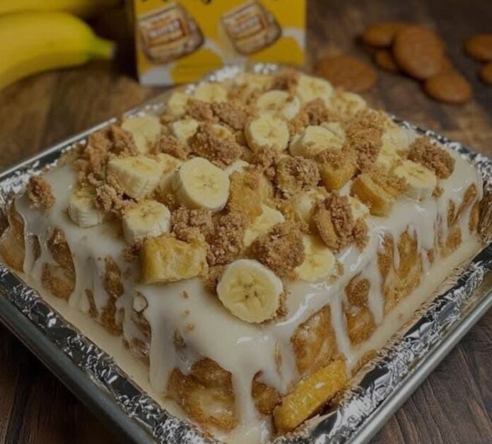 A square baking dish filled with Honey Bun Banana Pudding Cake. The cake has layers of honey buns, banana slices, and a creamy white pudding, topped with more banana slices and crumbled honey buns. A box of vanilla wafer cookies and a bunch of bananas are visible in the blurred background.