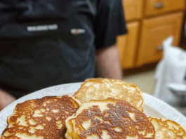 Old-Fashioned Pancakes A white plate filled with a stack of golden-brown pancakes, some dusted with powdered sugar. A person's torso is visible in the blurred background.