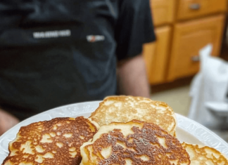 Old-Fashioned Pancakes A white plate filled with a stack of golden-brown pancakes, some dusted with powdered sugar. A person's torso is visible in the blurred background.