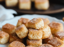 Cinnamon Sugar Biscuit Bites Close-up of a white plate filled with small, square Cinnamon Sugar Biscuit Bites. The bites are golden-brown and coated in cinnamon sugar. A white cloth is visible in the background.