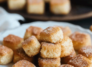 Close-up of a white plate filled with small, square Cinnamon Sugar Biscuit Bites. The bites are golden-brown and coated in cinnamon sugar. A white cloth is visible in the background.