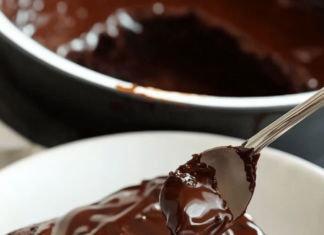 A square slice of dark chocolate zucchini cake on a white plate, topped with a glossy chocolate ganache. A spoonful of ganache is being drizzled over the cake. A black pot with more ganache is visible in the background.