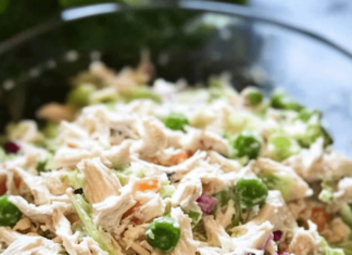 A close-up shot of a clear glass bowl filled with deli-style chicken salad. The salad includes shredded chicken, mayonnaise, chopped celery, red onion, and green peas.