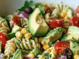 Avocado Pasta Salad Close-up of a white bowl filled with avocado pasta salad. The salad features rotini pasta, avocado chunks, halved grape tomatoes, corn kernels, red onion slices, black olives, and chopped parsley.