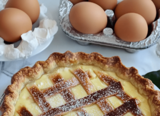 A partially sliced ricotta pie on a white plate. The pie has a golden brown crust, a creamy white filling, and a lattice design on top with powdered sugar. A carton of eggs is visible in the upper left corner.